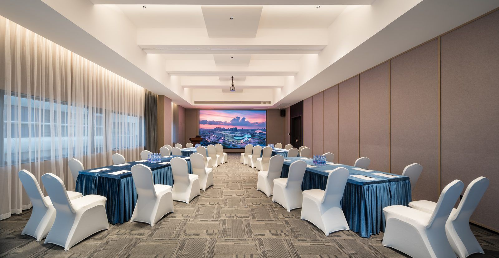 A conference room with tables and chairs, a large screen, and a view of a cityscape.