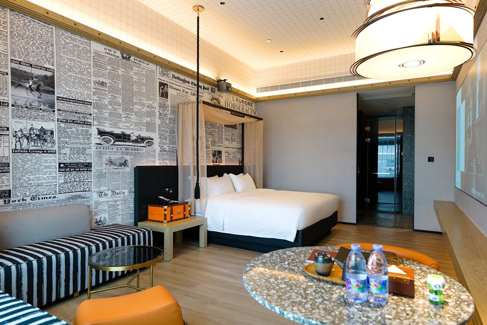 A hotel room with a bed, a seating area with a table, and a wall covered with newspaper clippings.