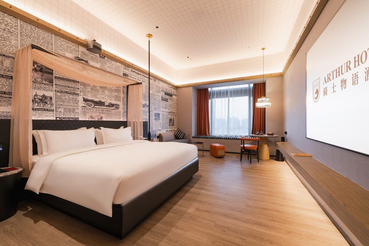 Habitación de hotel con una cama grande, un escritorio y una pantalla que muestra el nombre del hotel.