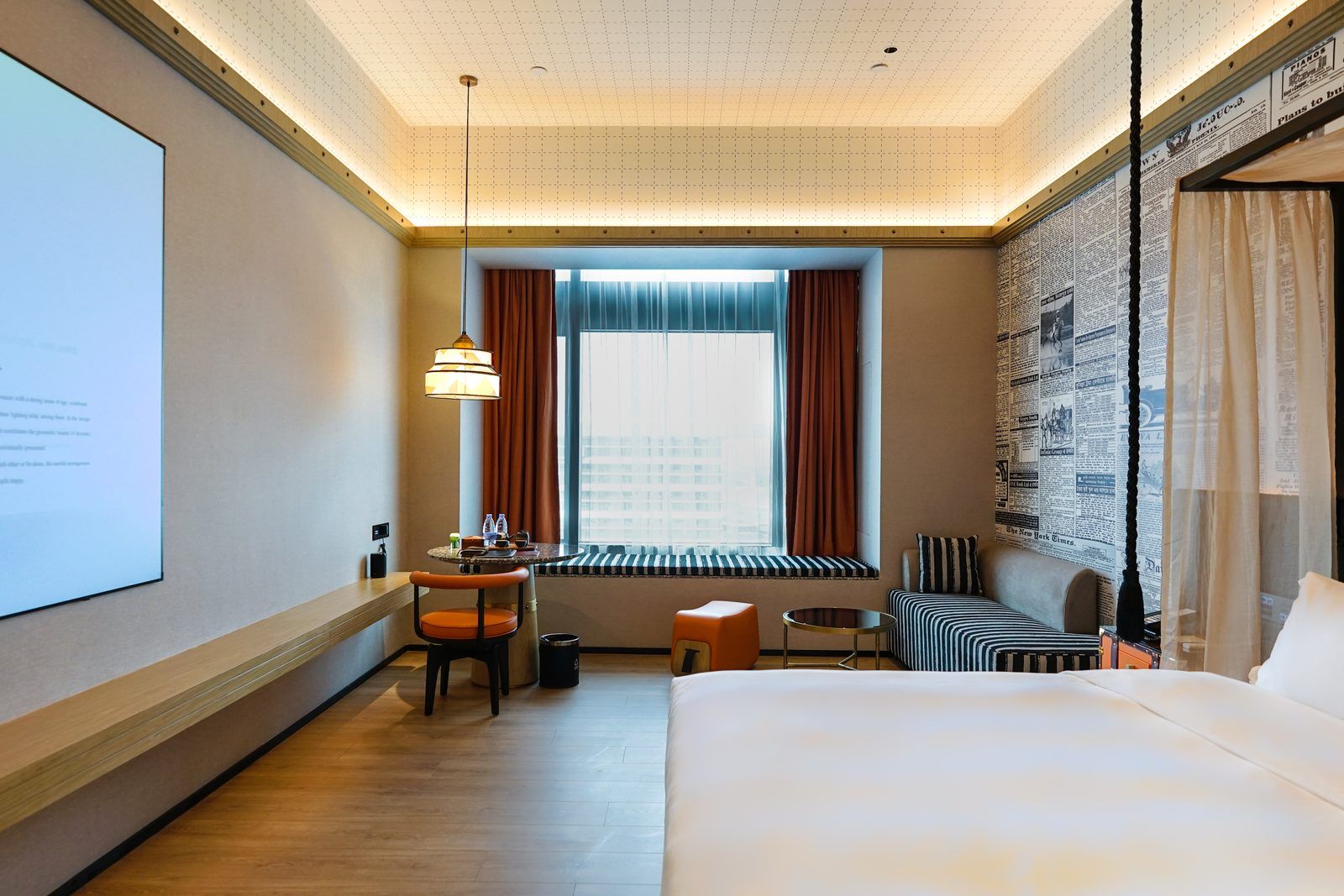 A hotel room with a bed, a desk with a chair, a window with curtains, and a wall with newspaper clippings.