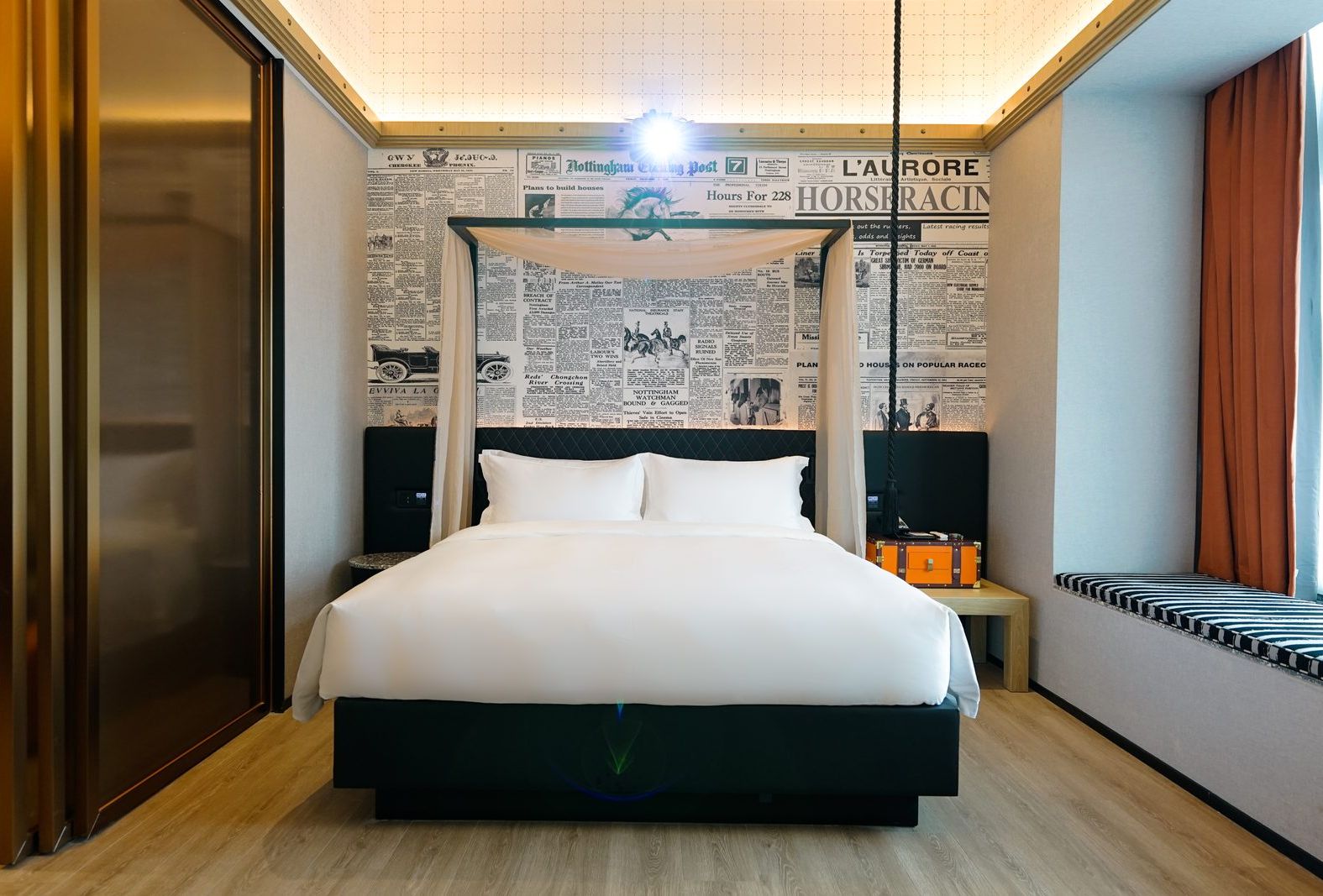 A hotel room with a large bed, a headboard with a canopy, a wall with newspaper clippings, a nightstand, and a suitcase.