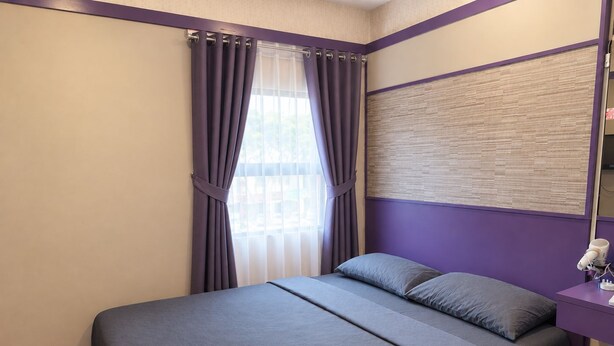 A bedroom with a bed, pillows, a window with curtains, a bedside table with a lamp, and a towel on the bed.