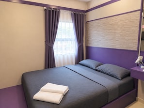 Un dormitorio con cama, almohadas, una ventana con cortinas, una mesita de noche con lámpara y una toalla sobre la cama.