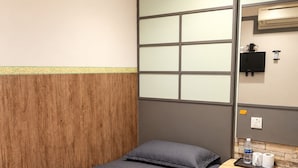 A single bed with a grey bedspread and pillows, a wooden headboard, a small bedside table with a water bottle and tissues, and a wall-mounted air conditioning unit.