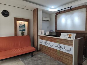 A hotel reception desk with a wooden finish, a clock on the wall, and a reception area with a sofa. - The Ardens Hotel - Taman Sri Yaacob (Johor Bahru)