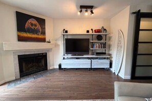 A living room with a fireplace, a flat-screen TV, and a bookshelf. - Tofino Surf Retreat — A Cozy Coastal Escape Near Tonquin Beach for 90+ day stays (Tofino)