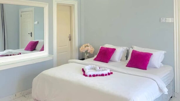 Un dormitorio con cama, espejo, puerta y una mesita con un florero.