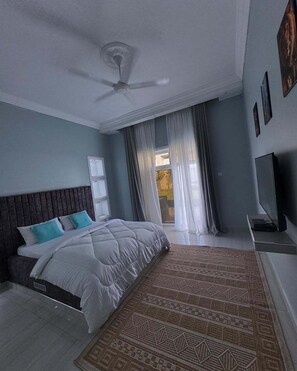 A bedroom with a bed, a ceiling fan, a TV, and a window with curtains. - Sunset Village Apartment Bagamoyo (Bagamoyo)