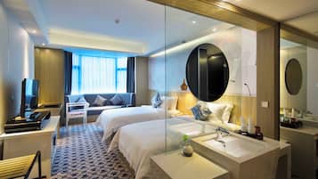 A hotel room with two beds, a desk, a chair, a television, and a bathroom with a mirror and sink.