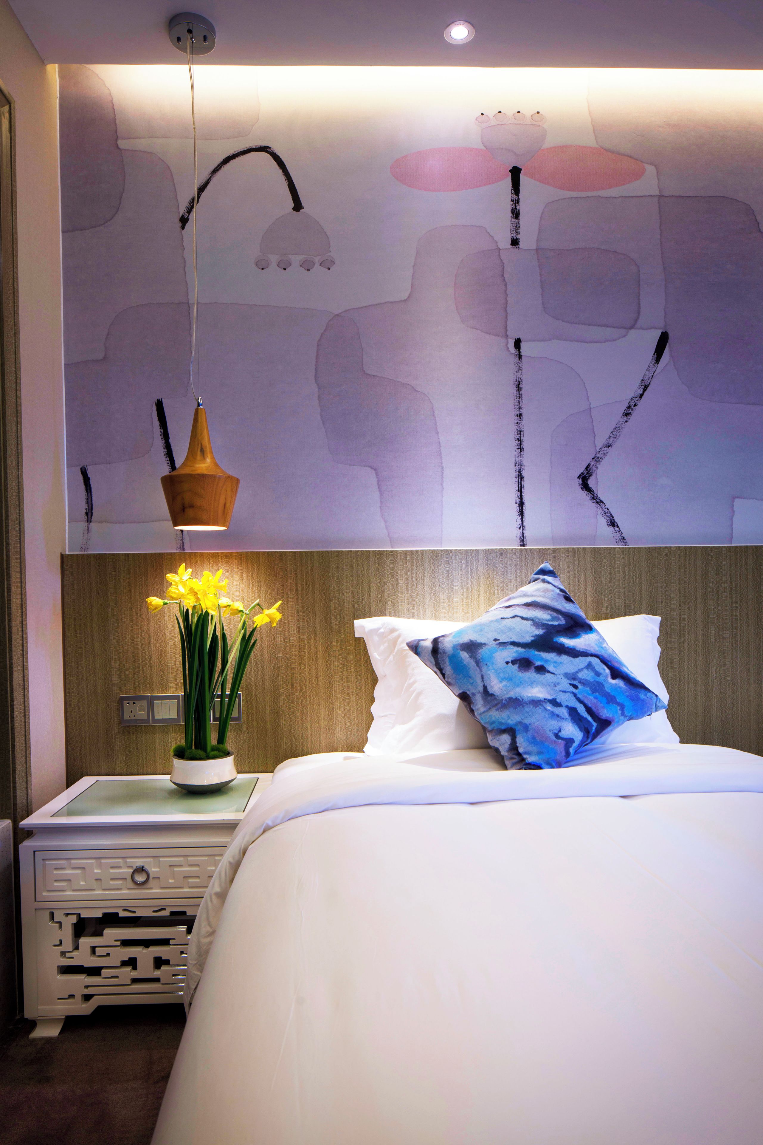 A hotel room with a bed, a nightstand, a vase with flowers, and a wall with abstract art.
