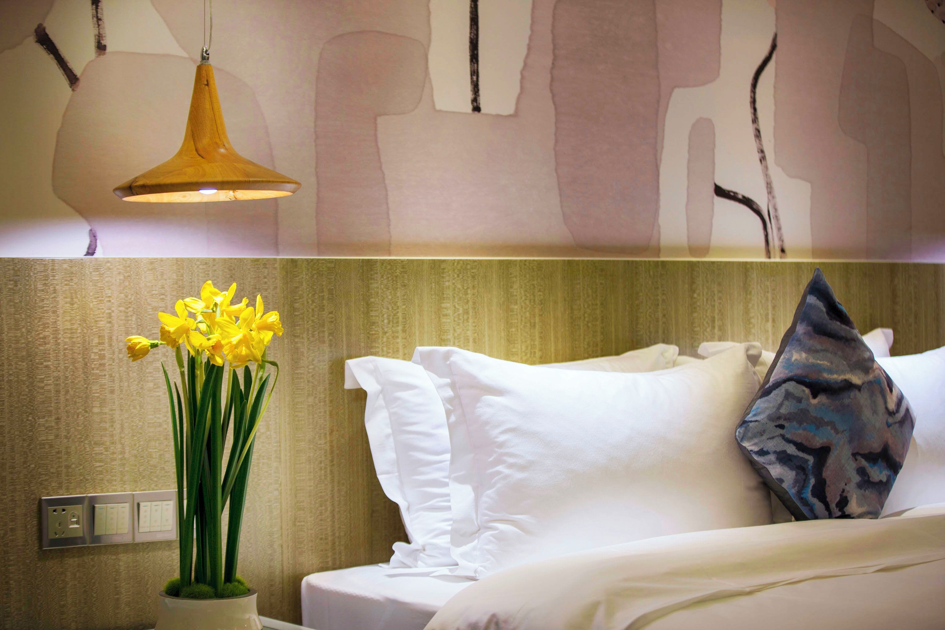 A hotel room with a bed, white pillows, a vase of yellow flowers, a wall-mounted light fixture, and a switchboard.