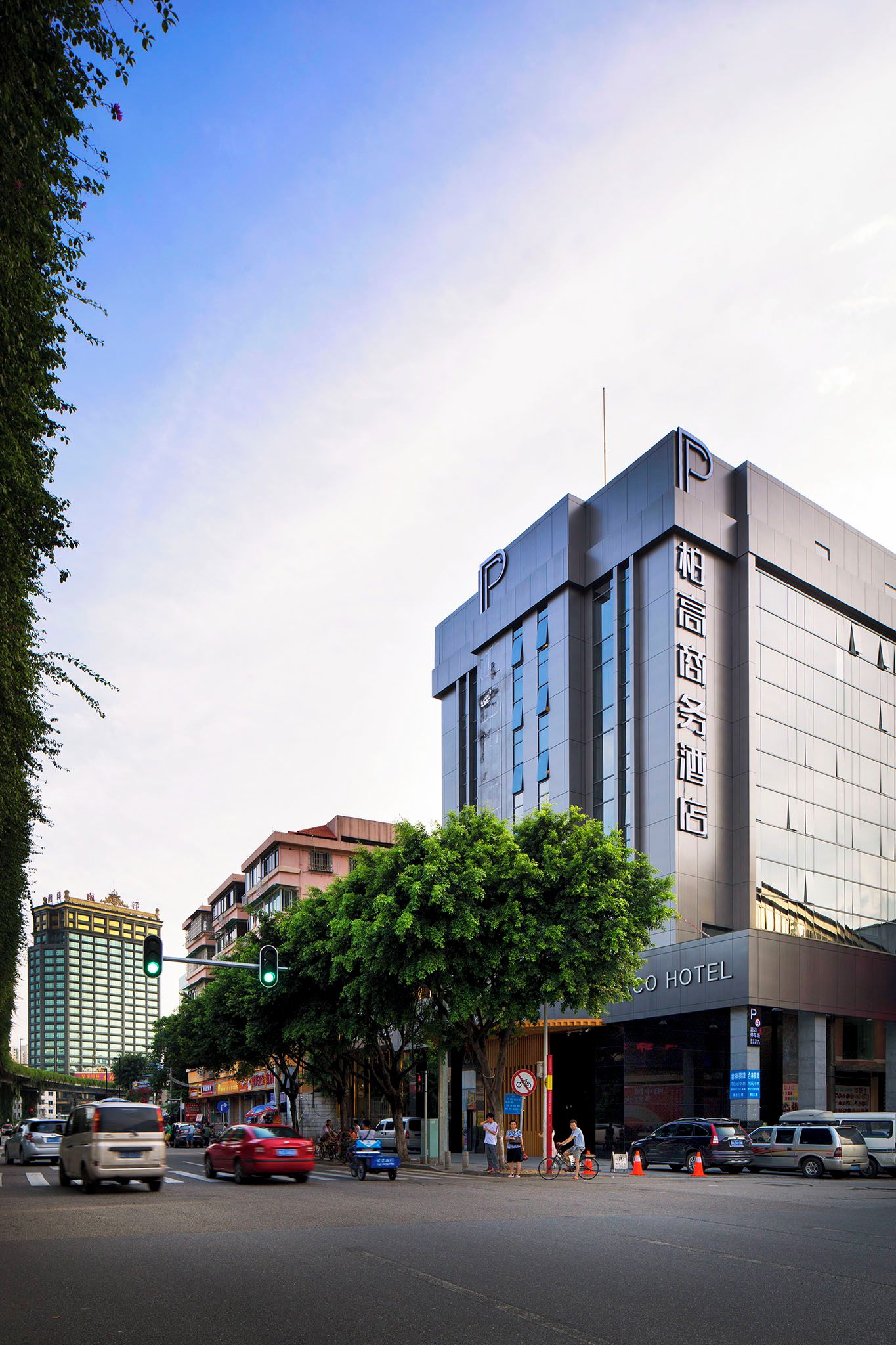 Edifício moderno de hotel com um logotipo 'P' em destaque, entrada visível e vista da rua com carros e pedestres.