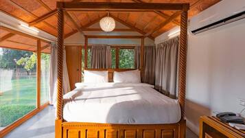 Una cama con dosel de madera y ropa de cama blanca, un ventilador de techo y vistas a un paisaje verde a través de amplios ventanales.
