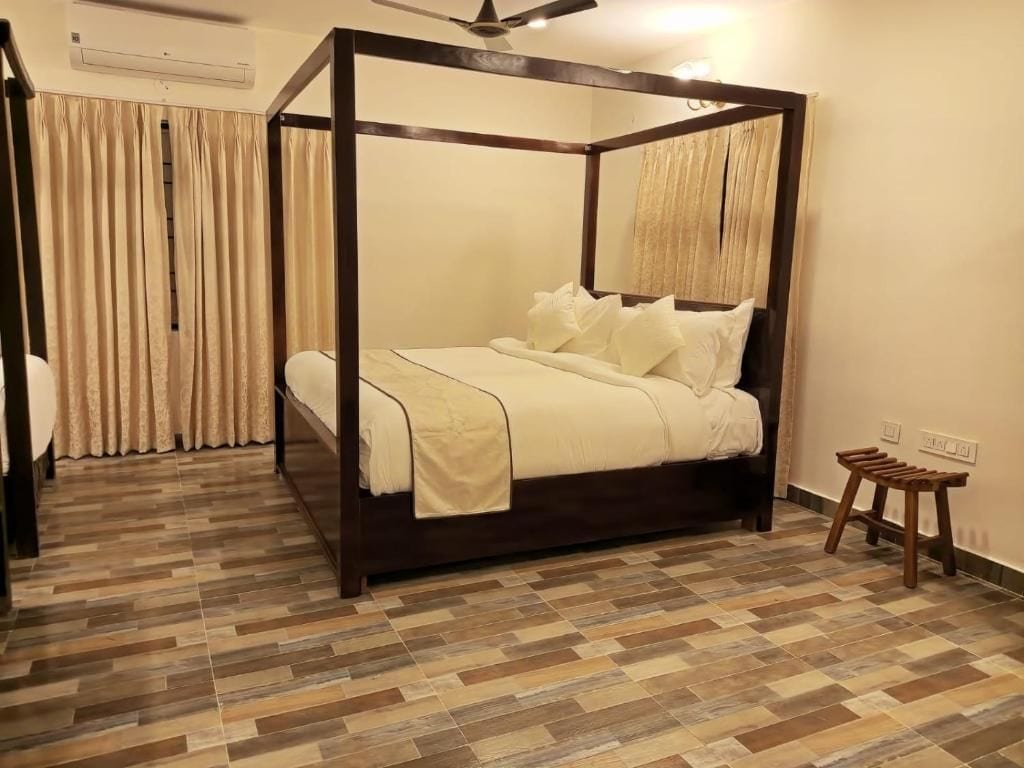 A four-poster bed with a wooden frame and white bedding, a wooden floor with a herringbone pattern, a ceiling fan, and a small wooden stool.