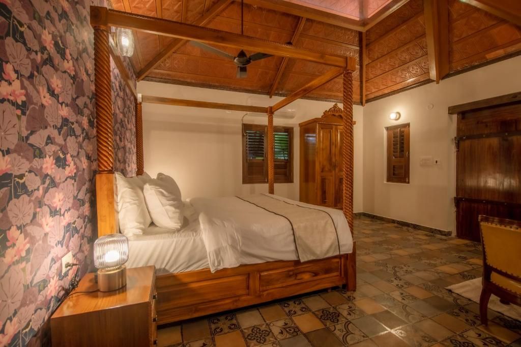 A four-poster bed with a wooden frame and white bedding, a nightstand with a lamp, and a chair in a room with patterned wallpaper and tiled flooring.