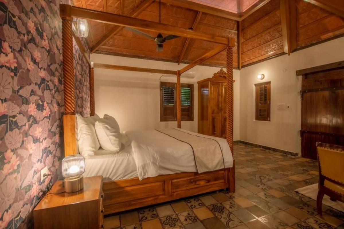 Una cama con dosel de madera y ropa de cama blanca, una mesita de noche con lámpara, y una silla en una habitación con papel tapiz estampado y suelo de baldosas.
