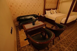 A bedroom with a four-poster bed, a nightstand, a wooden table with a vase of flowers, two leather chairs, and patterned wallpaper. - Brown Town Resort Spa and Convention  (Hyderabad)