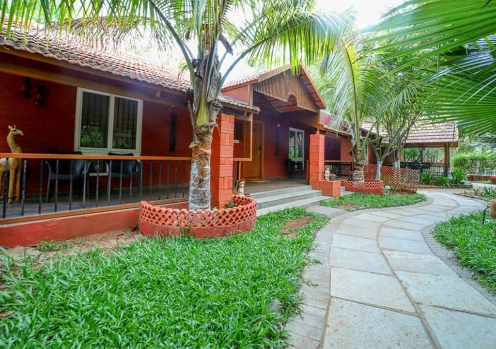 A red brick house with a tiled roof, a paved pathway, and a small garden area with grass and palm trees.