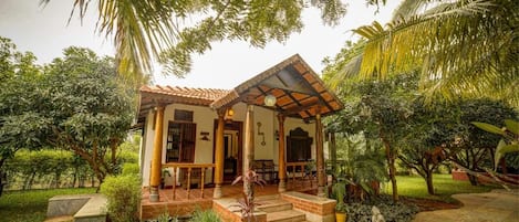 Una casa con techo de tejas, un porche con columnas de madera y un jardín con diversas plantas y una estatua.