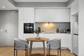 A modern kitchen with white cabinetry, a built-in oven, a microwave, and a dining area with two chairs and a round table. - Elevated 1-Bed Apartment Near Melbourne CBD (South Melbourne)