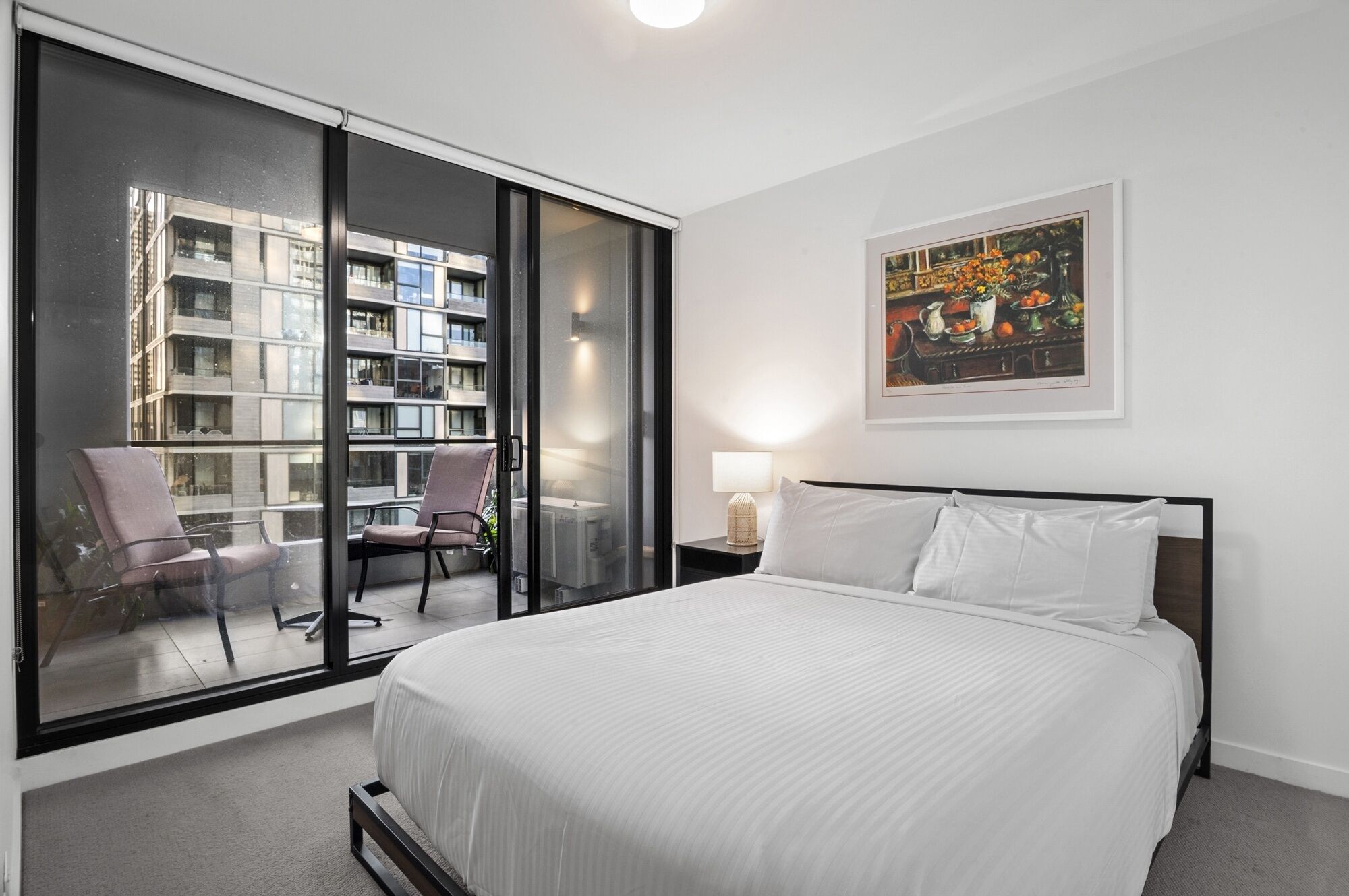 A hotel room with a large bed, a chair, a nightstand with a lamp, a painting on the wall, and a view of a building through a glass door.
