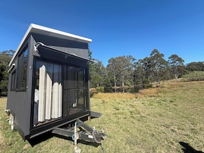 Una cabaña pequeña y moderna, negra, con puerta corredera de vidrio, ubicada en un campo cubierto de hierba con árboles al fondo.