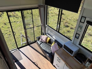 A room with a bed, a small table, a microwave, and a view of a grassy field through large windows.