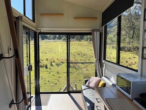 Una habitación con un amplio ventanal corredera que da a un campo cubierto de césped, un sofá con cojines y un microondas en la encimera.