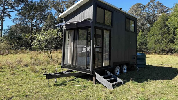 A modern, compact mobile home with a large sliding door and a small porch area, situated in a grassy field with trees in the background. - Hamilton Country Retreat 1 by Tiny Away (Kangaroo Valley)
