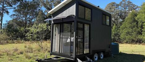 Une maison mobile moderne et compacte, dotée d’une grande porte coulissante et d’un petit espace porche, située dans un champ verdoyant avec des arbres en arrière-plan.