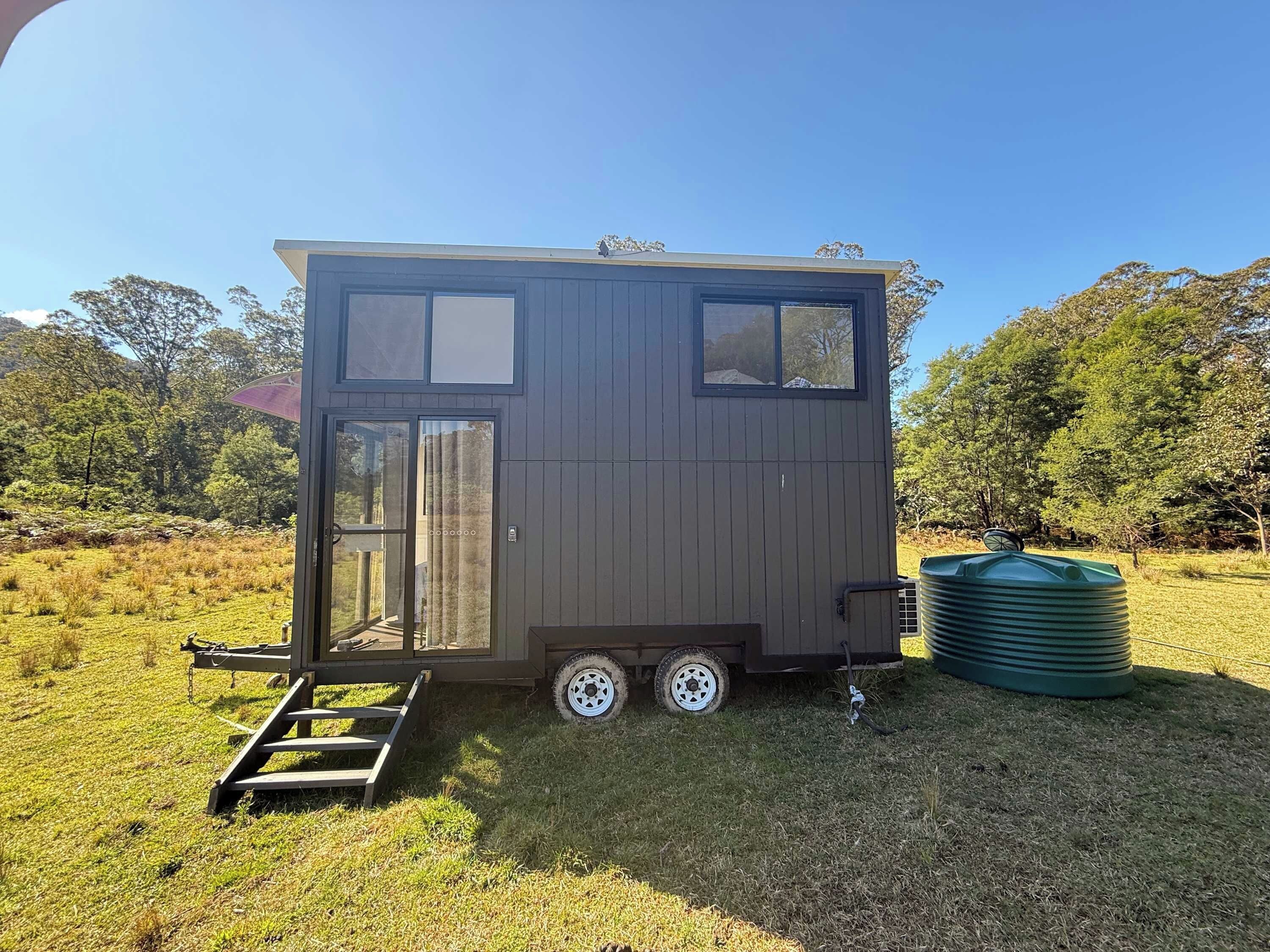 A small, two-story mobile home with a sliding door and a window, situated in a grassy field with trees in the background.