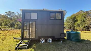 A small, two-story mobile home with a sliding door and a window, situated in a grassy field with trees in the background.