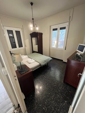 Ein Schlafzimmer mit einem Bett, einem Schreibtisch mit Computer, einem Spiegel und zwei Fenstern.