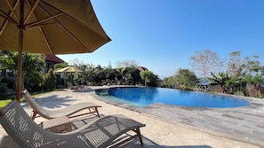 Una piscina con sillas de descanso y sombrillas, rodeada de follaje y un cielo despejado.
