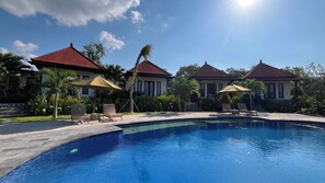 A swimming pool with lounge chairs and umbrellas, surrounded by palm trees and a building with a red roof. - The Hills Penida (Klungkung)