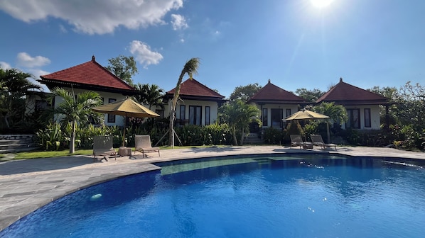 A swimming pool with lounge chairs and umbrellas, surrounded by palm trees and a building with a red roof.