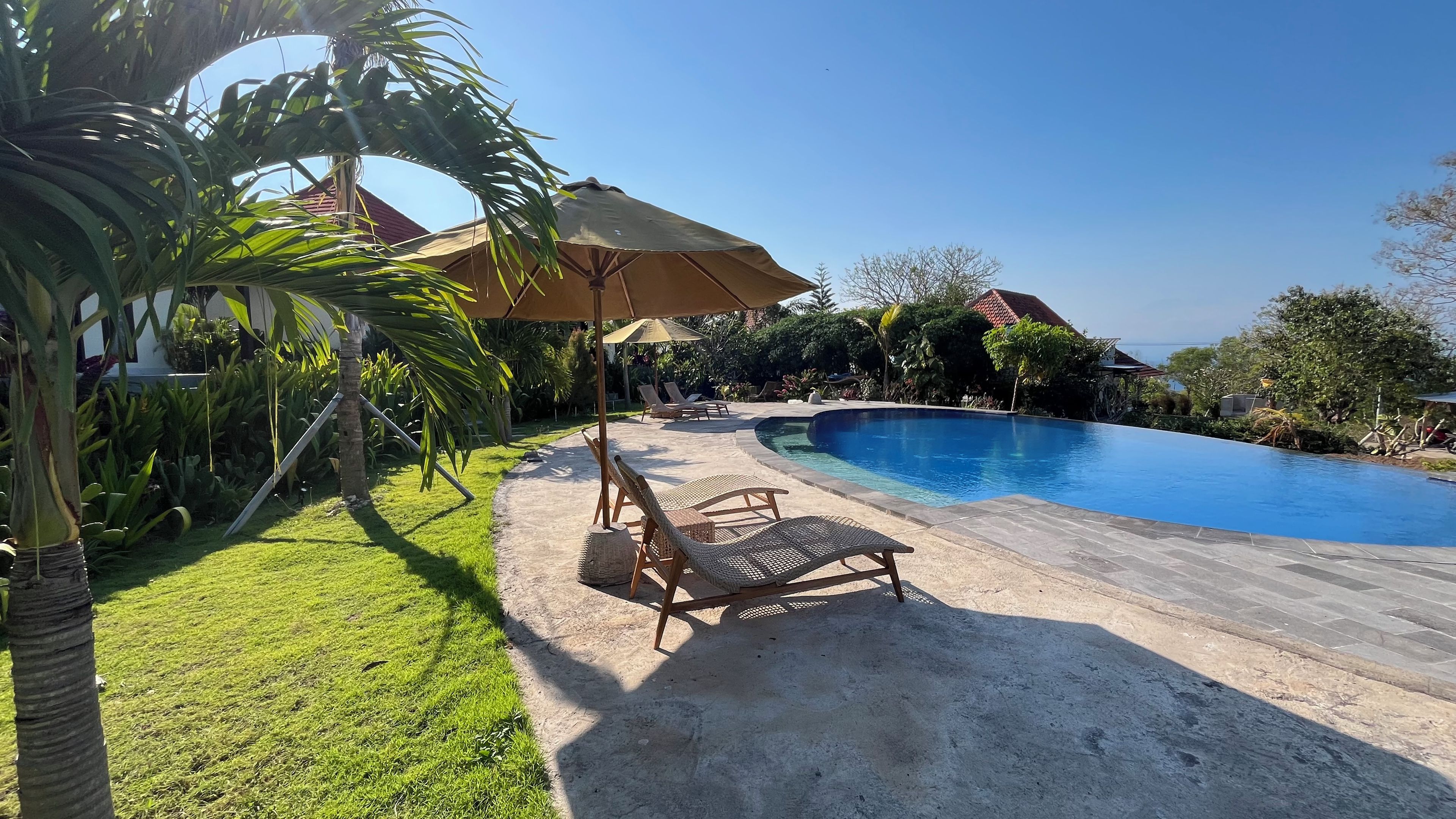 Una piscina con sillones de descanso y sombrillas, rodeada de vegetación y un cielo despejado.