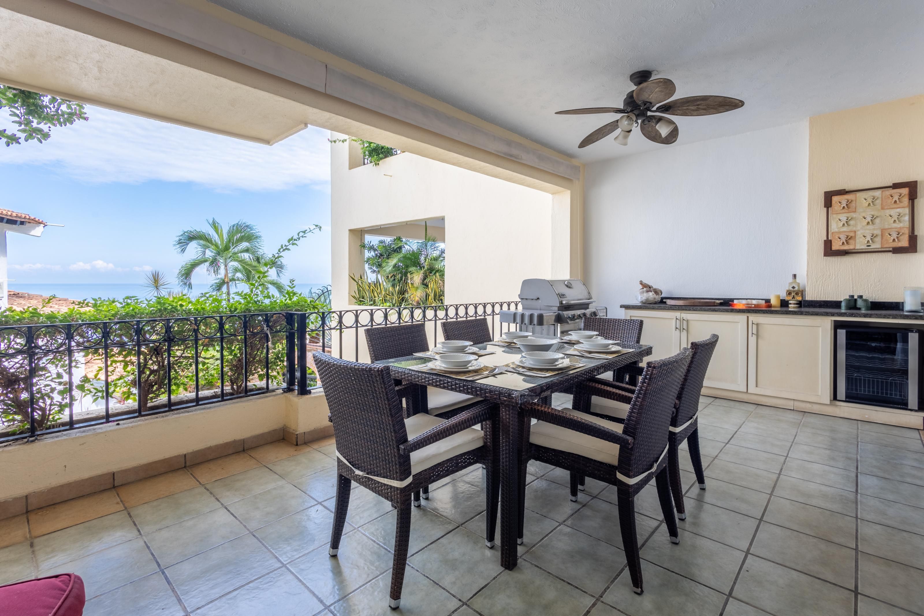 A balcony with a dining table set for four, a ceiling fan, and a kitchen area with a grill.