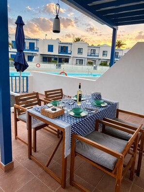 Eine Terrasse mit Esstisch, mit Blick auf einen Swimmingpool und Gebäude mit blauen Akzenten.