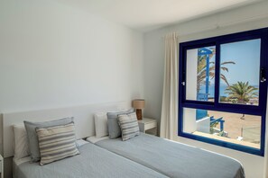 A bedroom with a bed, two pillows, a nightstand, a lamp, and a window with a view of palm trees and a beach. - Casa Alejandra, 2 bedrooms, 2 bathrooms and shared pool in Playa Blanca (Playa Blanca)