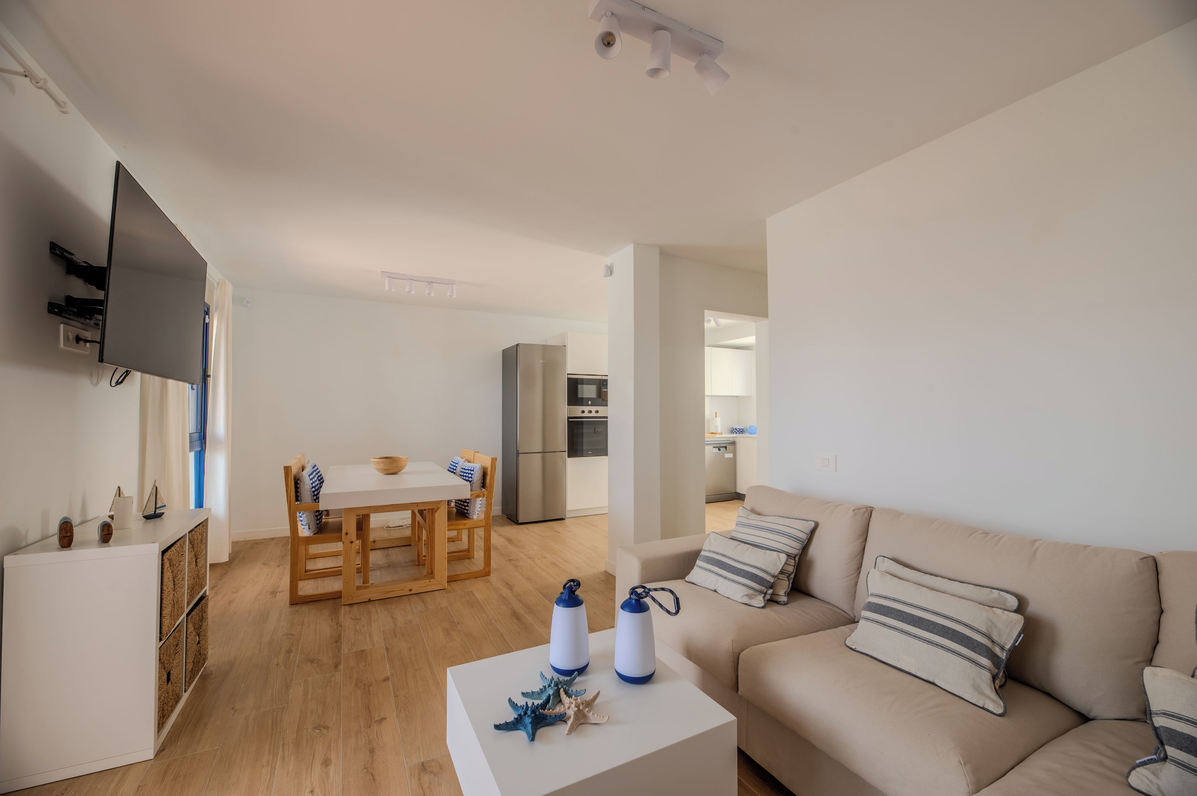 Un salon moderne comprenant un canapé, une table à manger avec des chaises, un coin cuisine avec un réfrigérateur et un téléviseur à écran plat fixé au mur.
