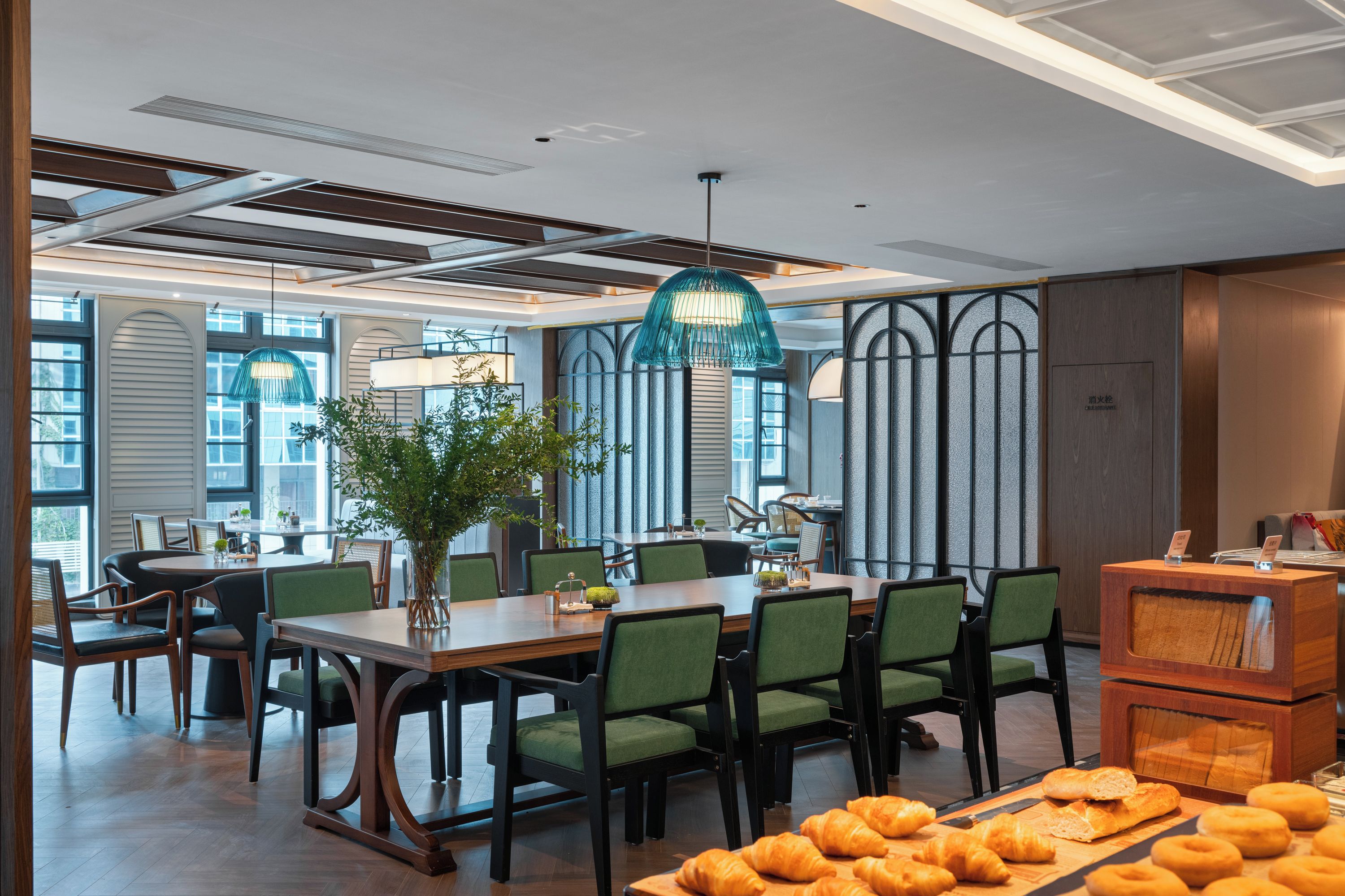 Un comedor moderno con mesas y sillas de madera, una lámpara colgante grande y vistas de la ciudad a través de amplios ventanales.