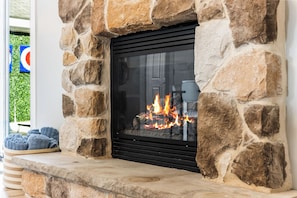 A stone fireplace with a glass door, a fire burning inside, and a stone bench with cushions to the left. - NEW! Indoor Lounge Pool Hot tub Glamping Karaoke (Tobyhanna)