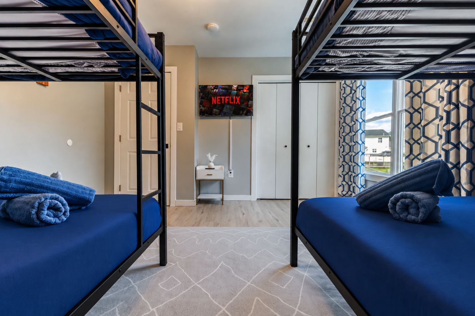 Une chambre avec des lits superposés, un bureau, une chaise, un téléviseur affichant le logo de Netflix et une fenêtre avec des rideaux à motifs.