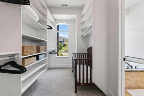 Ein schmaler Flur mit weißen Regalen, einem hölzernen Babybett und Blick auf ein Haus durch ein Fenster.