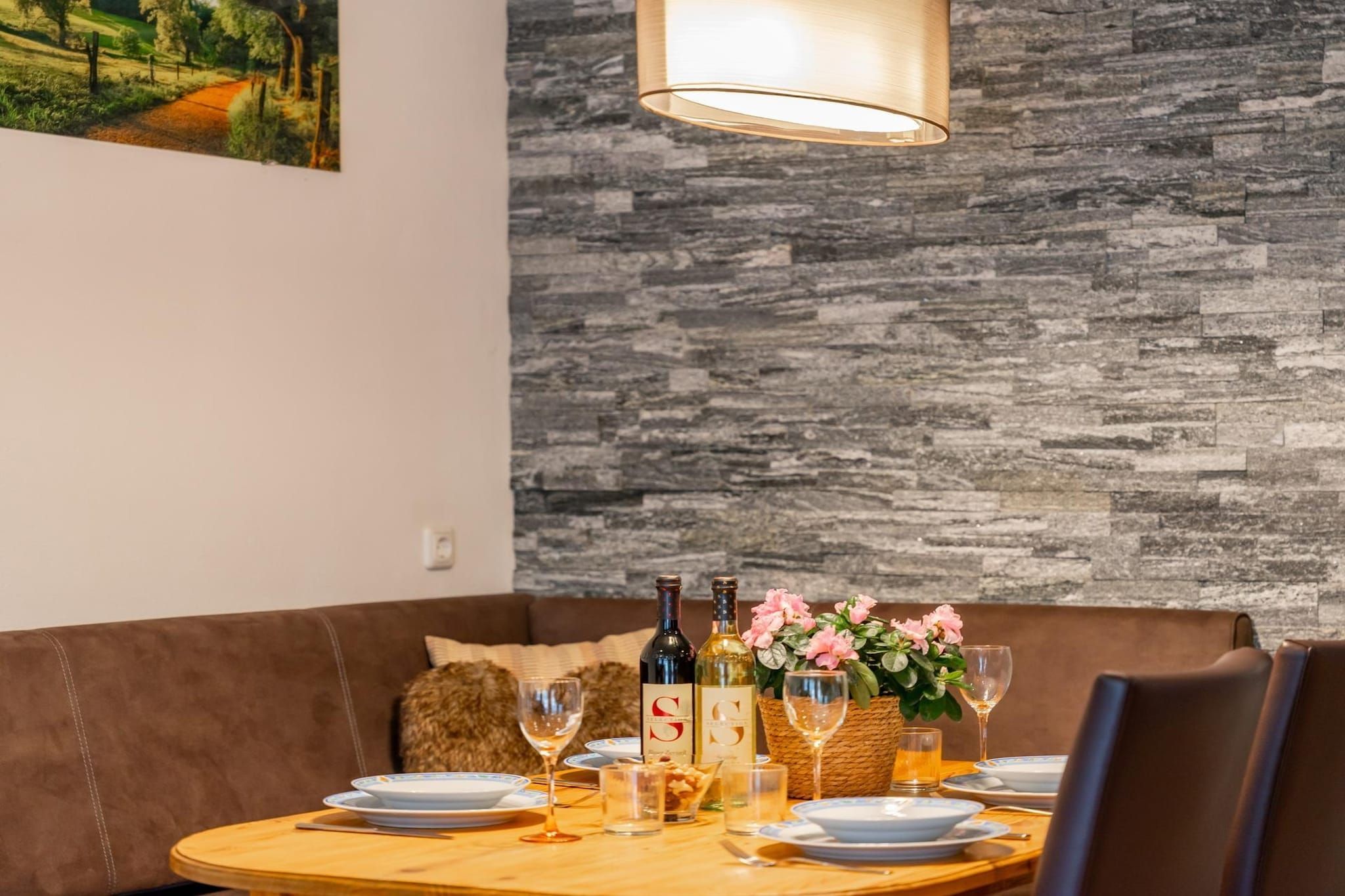 Spiseområde med et rundt bord dækket til to personer, vinflasker, glas og en kurv med blomster.