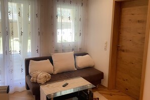 Ein gemütliches Wohnzimmer mit einem braunen Sofa, zwei weißen Kissen, einer flauschigen Decke und einem Glastisch mit einer Fernbedienung.