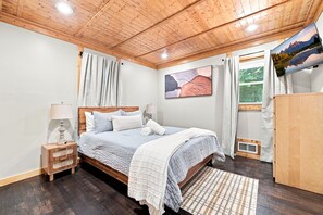 Un dormitorio con techo de madera, una cama con colcha blanca y azul, una mesita de noche, una lámpara, un cuadro y una ventana con cortinas.