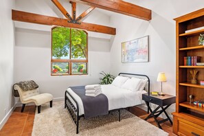 Quarto com cama, cadeira, mesa de cabeceira, luminária, estante e janela com vista para o verde.