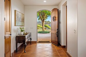 Corredor com porta de madeira que dá para área externa com árvore e vista de vinhedo.